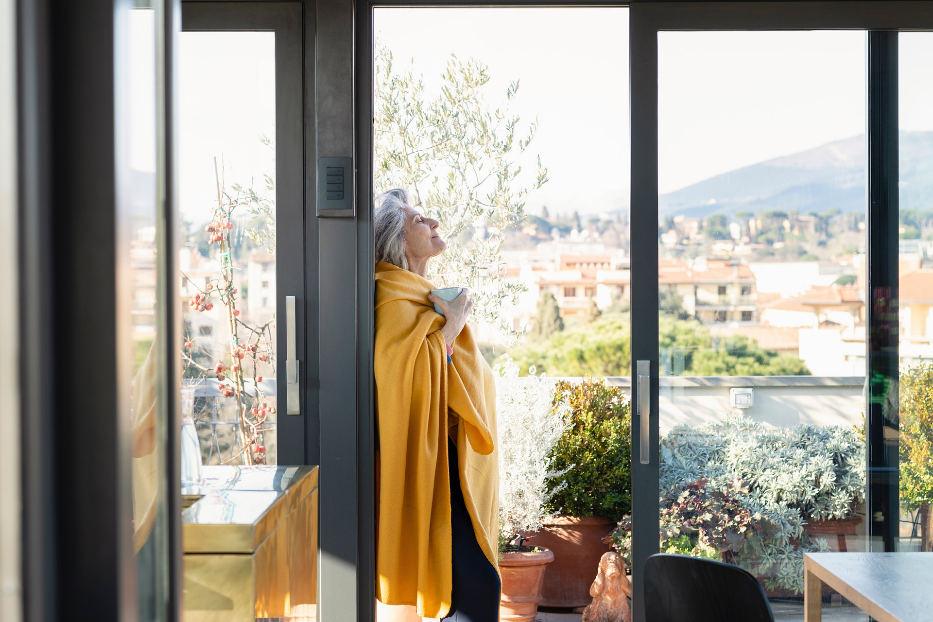 ältere Dame mit Tasse in der Hand lehnt an Balkontür