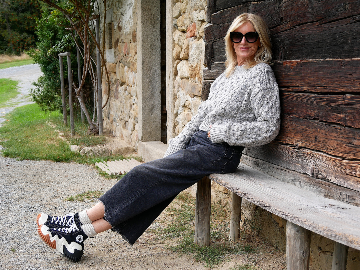Tina Artz mit Sonnenbrille sitzt auf einer Bank vor einem ländlichen Gebäude