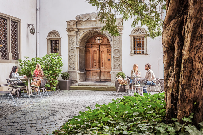 Reisen kennt kein Alter Bodensee Reiseführer für Senioren: Vier Personen sitzen im idyllischen Innenhof des Rosengartenmuseums an Tischen und unterhalten sich