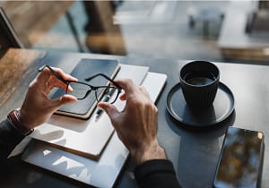 Person sitzt Tisch, auf dem Mobiltelefon, Kaffeetasse & Notizbücher liegen, und hält eine Brille in der Hand