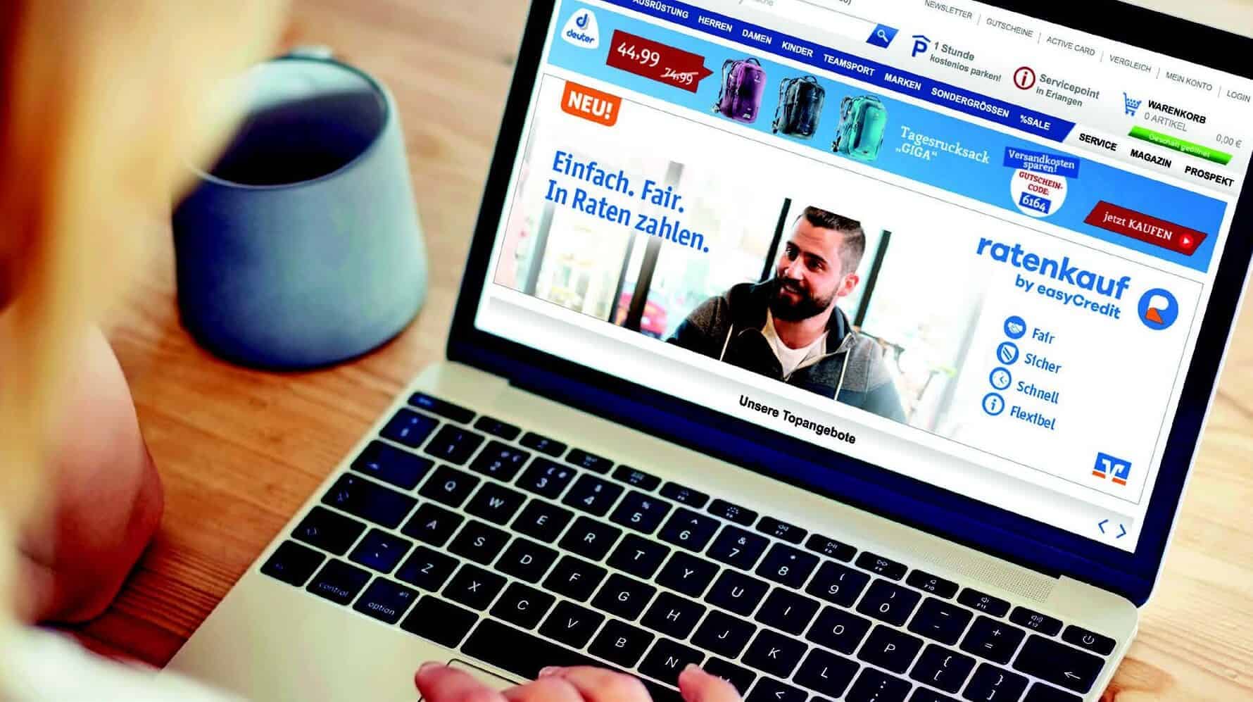 Blick über Schulter einer Person, die an Holztisch sitzt, eine blaue Tasse vor sich hat, und auf einem Laptop die Website von easyCredit-Ratenkauf anschaut.