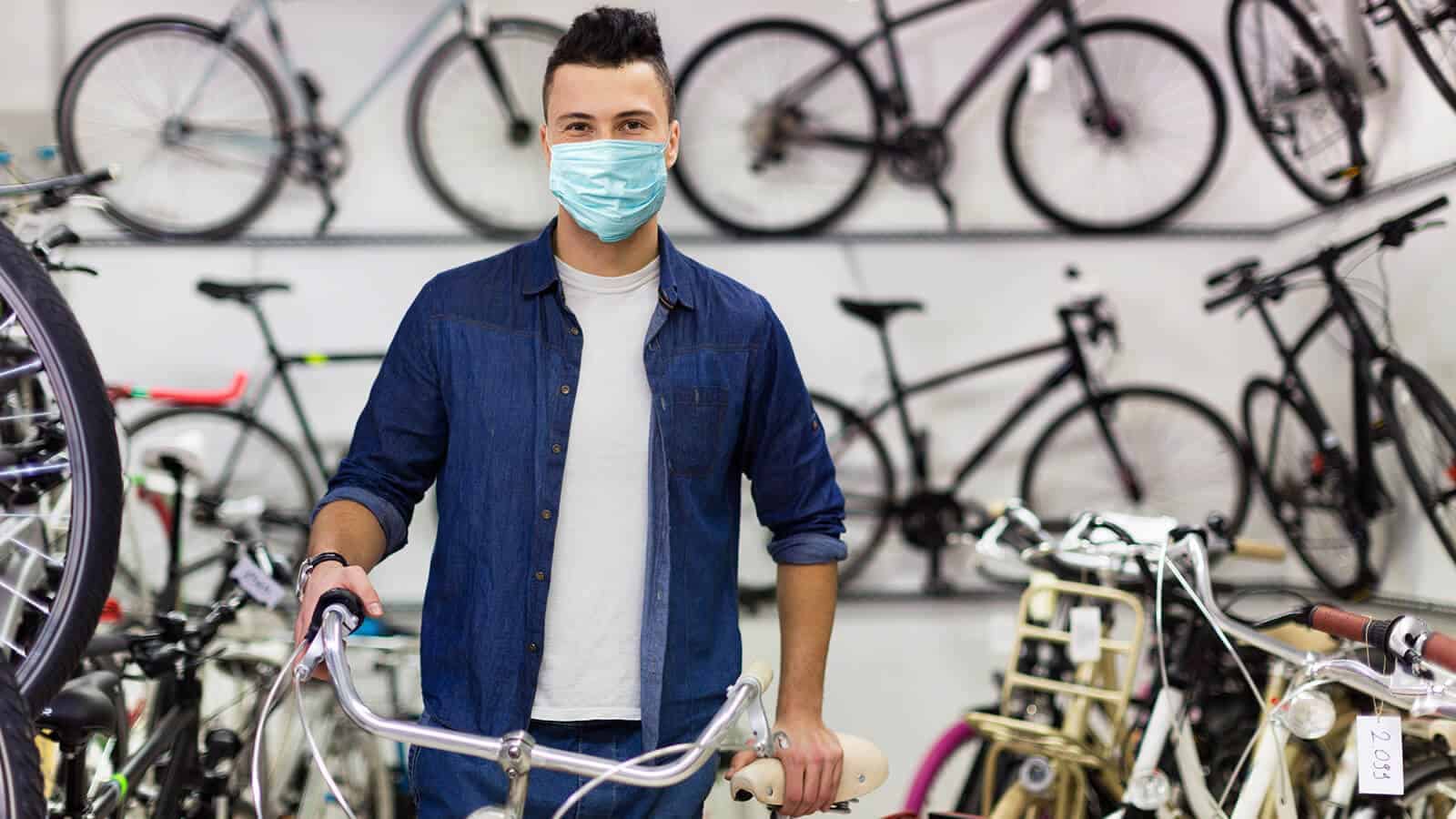 Junger Mann mit Mund-Nasen-Maske steht in Fahrradgeschäft und hält Fahrrad an Lenker fest.