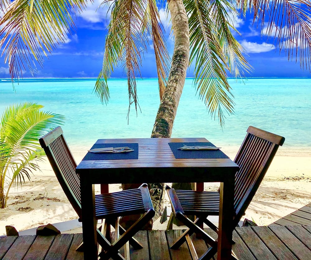 Tisch für zwei in Bora Bora. Im Hintergrund ist eine Palme, weißer Sandstrand und blaues Meer und Horizont erkennbar.