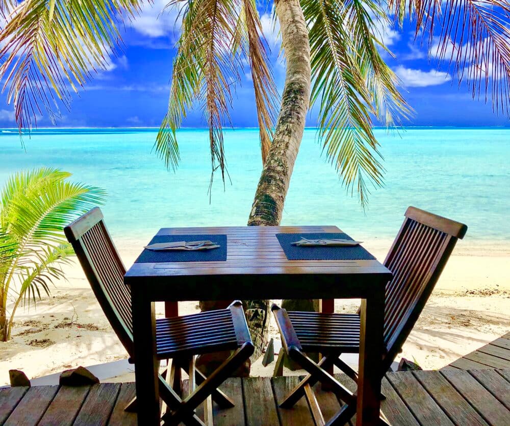 Tisch für zwei in Bora Bora. Im Hintergrund ist eine Palme, weißer Sandstrand und blaues Meer und Horizont erkennbar.