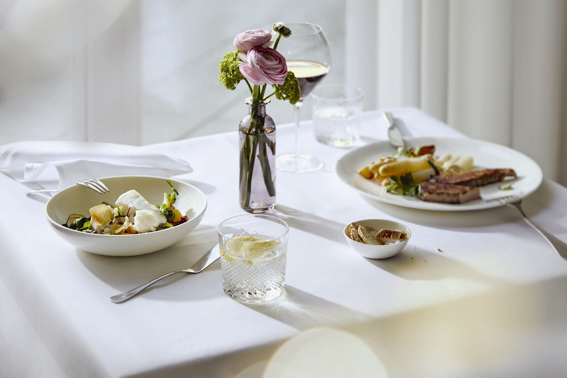 Ein gedeckter Tisch mit leckerem Essen, weißer Tischdecke und Blumen als Deko in der Tertianum Seniorereseidenz Bayern.