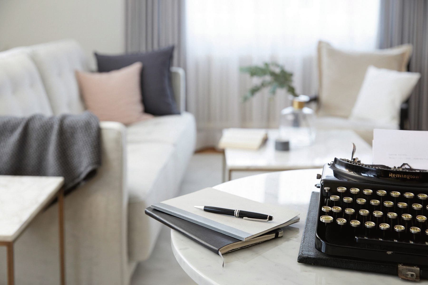 Tisch mit alter Schreibmaschine, zwei Notizheften und Stift, im Hintergrund ein Sofa, ein Sessel und zwei Beistelltische