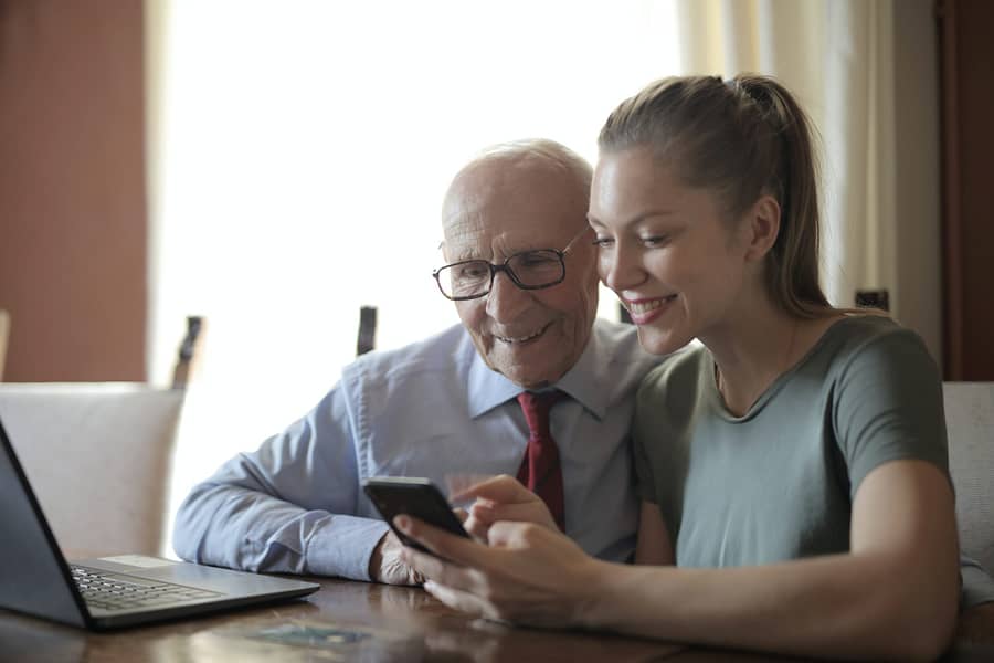 Ein Senior und eine junge Frau sitzen an einem Tisch und blicken zusammen auf ein Smartphone. Beide lächeln.