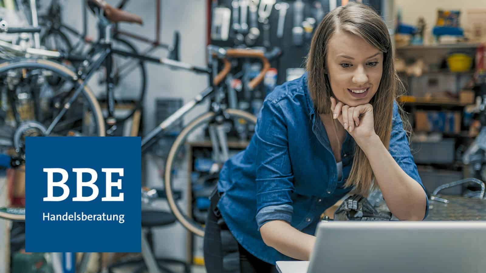 Mitarbeiterin von Fahrradladen arbeitet an Laptop. Links Logo der "BBE Handelsberatung"