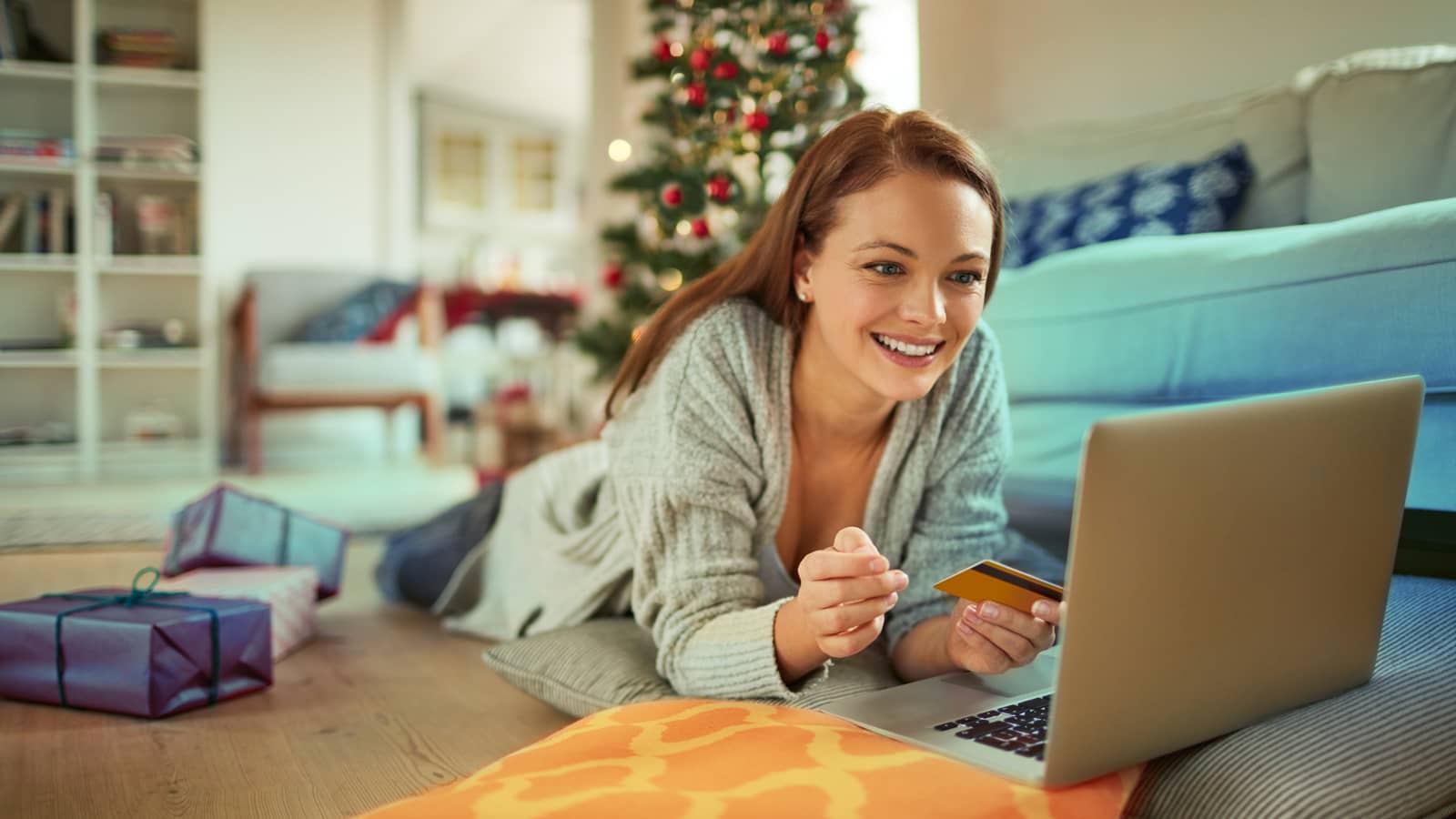 Glückliche Frau liegt mit Laptop im Wohnzimmer und hat Kreditkarte in der Hand.