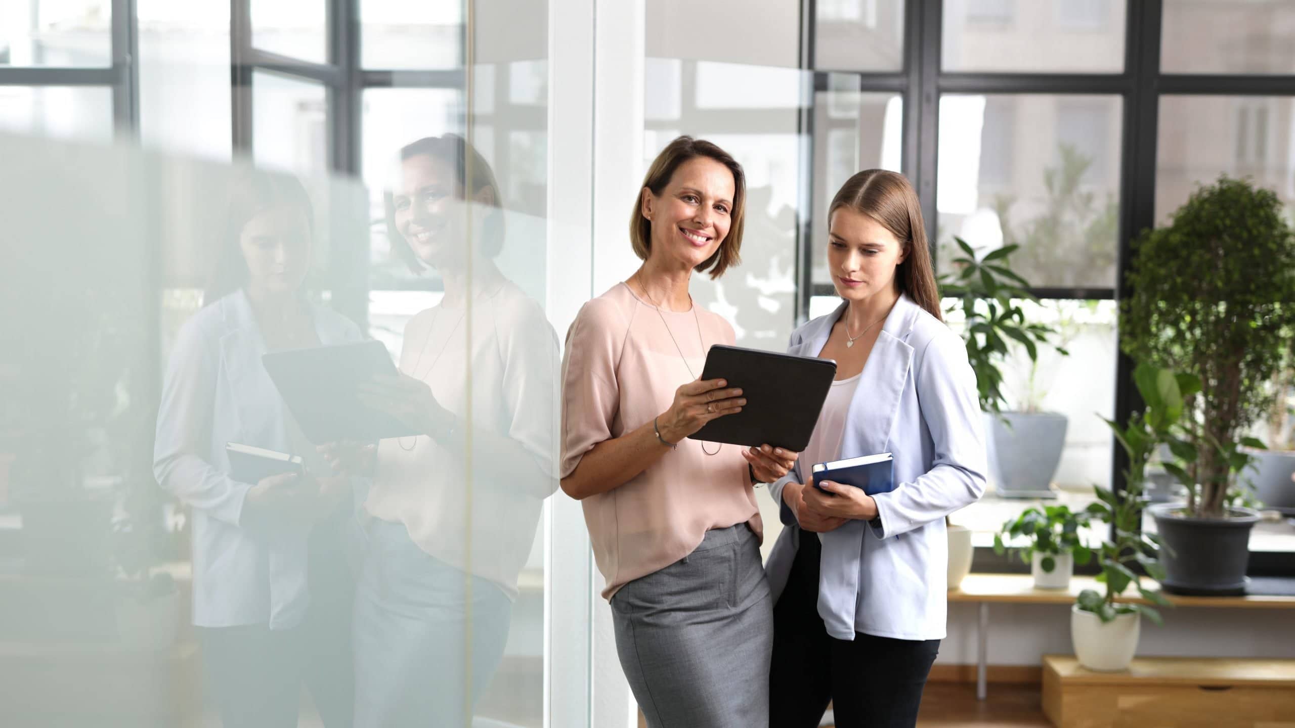 Zwei Frauen stehen in einem modernen Büro, lächeln und betrachten gemeinsam ein Tablet.
