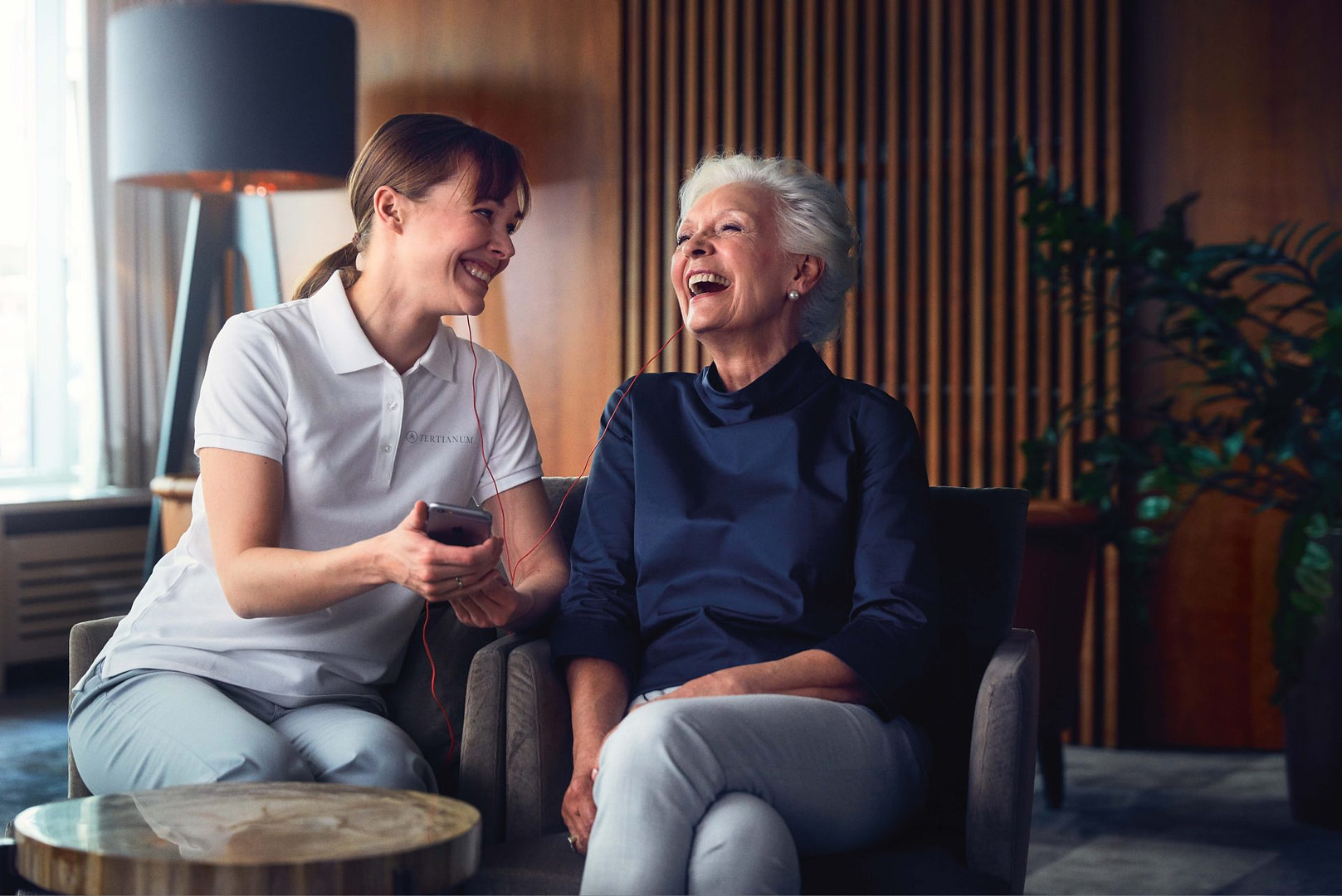 Eine Seniorin sitz neben einer Pflegerin, beide lachen fröhlich.