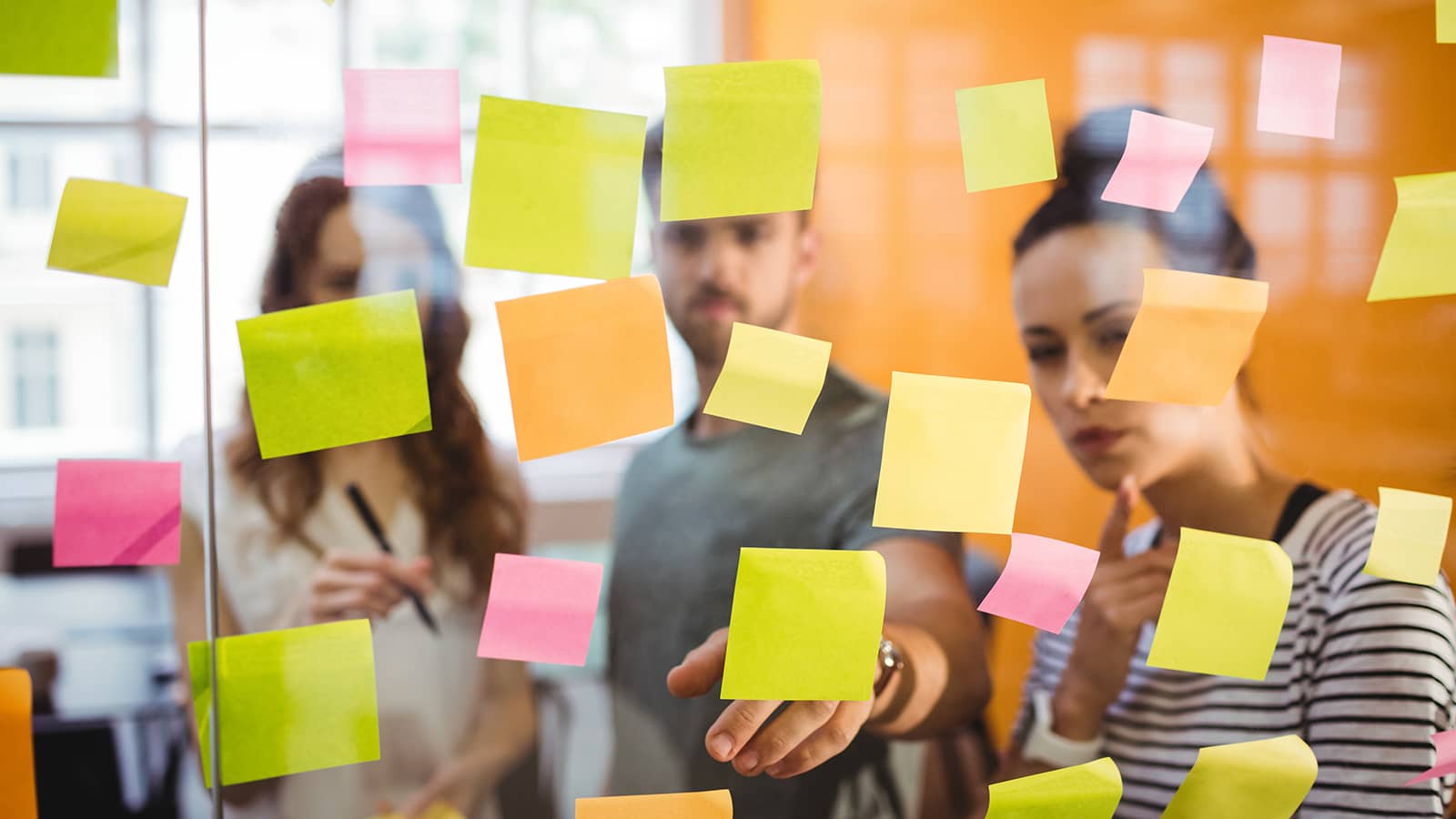 Trends erkennen, Zielgruppen ansprechen und Marketingstrategien anpassen - drei Personen stehen vor Glaswand und diskutieren Ideen auf Post-It Zetteln