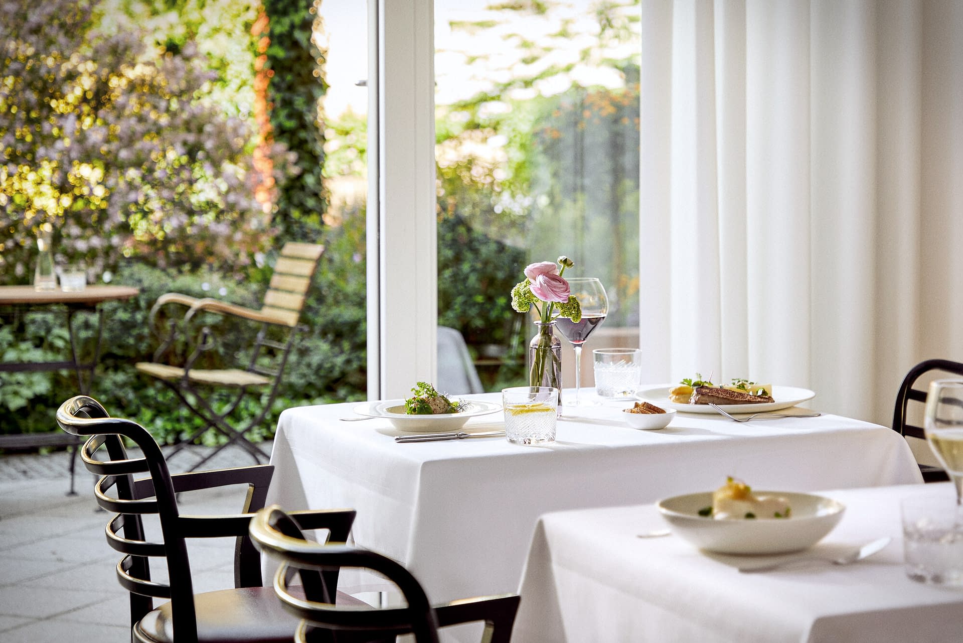 Ein weiß gedeckter Tisch mit gutem Essen und Blumen mit Blick in den grünen Garten in der Tertianum Senioreneinrichtung.
