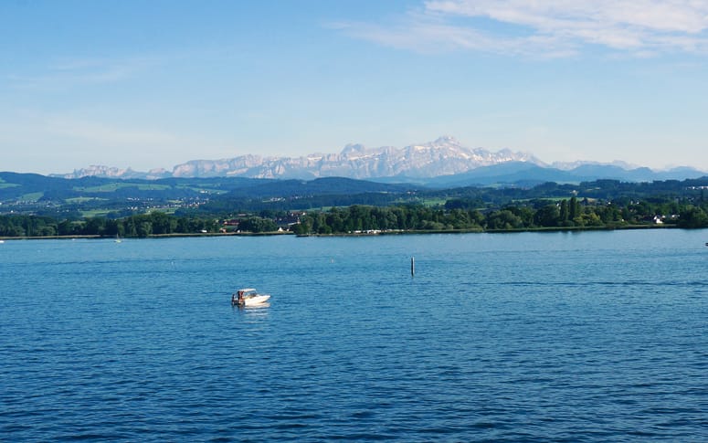Reisen kennt kein Alter Bodensee Reiseführer für Senioren: Blick über den Bodensee. im Vordergrund befindet sich ein kleines Boot, am Horizont sind die Gipfel der Alpen erkennbar